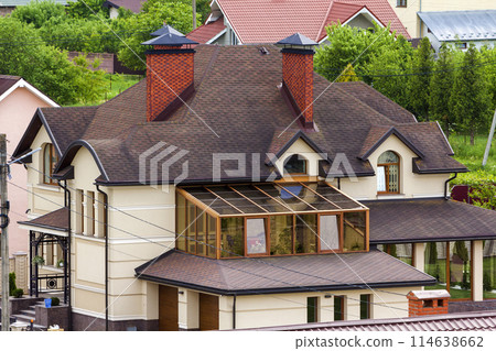 Close-up of big new comfortable two-storied new cottage with green tile roof, big yard, plastic windows and high chimneys in modern quiet suburban residential area. Perfect investment in dream house. 114638662