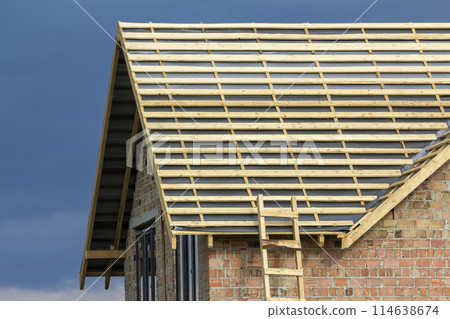 Close-up detail of new brick house top with two narrow plastic attic windows and wooden roof frame under construction on dark blue sky background. Real estate property and professional work concept. 114638674
