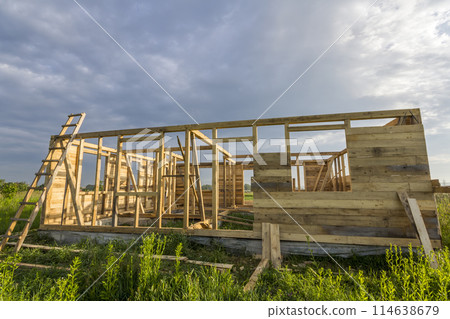 Unfinished ecological house under construction in green field on blue sky background. Trenches filled with cement and wooden walls with window openings of future comfortable cottage in quiet area. 114638679