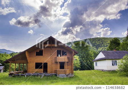 New wooden ecological cottage with balcony, terrace, steep roof from natural materials under construction on grassy meadow on forested hills background. Old traditions and modern building concept. 114638682