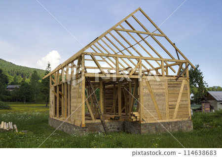 New wooden house under construction in quiet rural neighborhood. Timber frame of natural materials for walls and roof on stone foundation. Property, professional building and reconstruction concept. 114638683