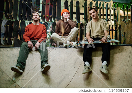 Group of self-confident young male and female skateboard riders in streetwear sitting relaxed in skatepark and looking at camera Group of self-confident young male and female skateboard riders in streetwear sitting relaxed in skatepark and looking at camera 114639397