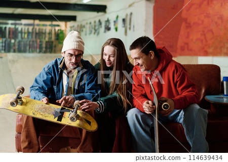 Two gen Z boys and girl in streetwear sitting on couch in skatepark discussing skateboard design, copy space 114639434
