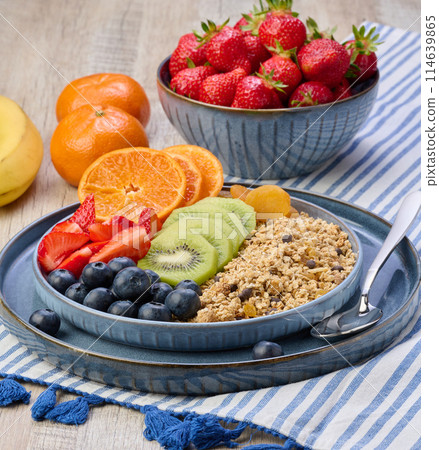 Granola with strawberries, kiwi, banana and blueberries in a round plate on a white table. 114639865