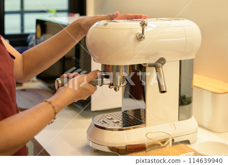 Female preparing fresh coffee while using modern coffee maker in the kitchen at home. Female preparing fresh coffee while using modern coffee maker in the kitchen at home. 114639940