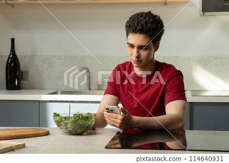 Young Man Sitting at Kitchen Table Looking at Cell Phone Young Man Sitting at Kitchen Table Looking at Cell Phone 114640931