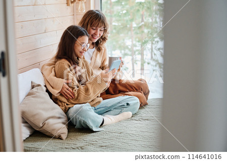 Caucasian woman and her daughter sitting relaxed on bed at home surfing Internet on smartphone, copy space Caucasian woman and her daughter sitting relaxed on bed at home surfing Internet on smartphone, copy space 114641016
