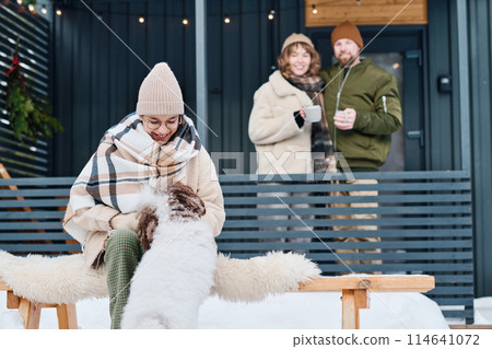 Joyful teen girl wearing warm clothes sitting outdoors spending time with her dog, happy parents standing porch in background 114641072