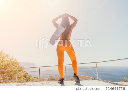 Fitness woman sea. Happy middle aged woman in orange sportswear exercises morning outdoors on yoga mat with laptop in park over ocean beach. Female fitness pilates yoga routine. Healthy lifestyle. Fitness woman sea. Happy middle aged woman in orange sportswear exercises morning outdoors on yoga mat with laptop in park over ocean beach. Female fitness pilates yoga routine. Healthy lifestyle. 114641756