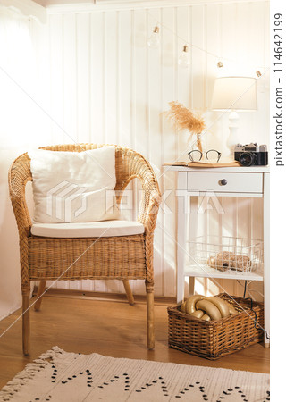 Wicker peacock chair with pillow and bedside table with book, retro camera, glasses and lamp in beige light wooden interior 114642199