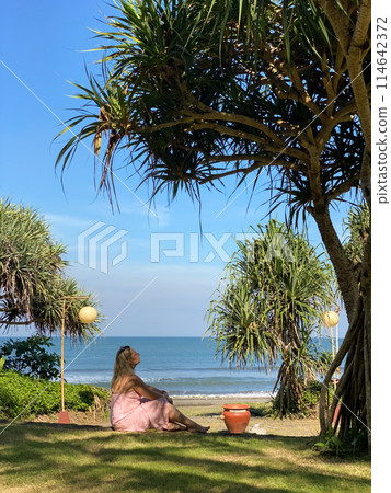 Woman in a red dress near a palm tree looking at the ocean Woman in a red dress near a palm tree looking at the ocean 114642372