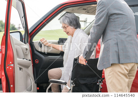 An elderly woman on a wheelchair after getting off the car and a man assisting (transfer) 114645540