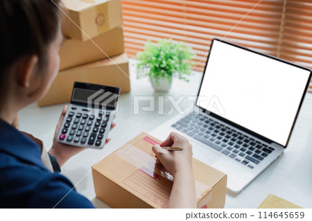 Entrepreneur using calculator in her hand, calculating financial expense at home office, online market packing box delivery Entrepreneur using calculator in her hand, calculating financial expense at home office, online market packing box delivery 114645659