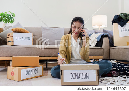 Young woman Asian of charity putting clothes in boxes with donations lettering and using smartphone to contact donations Young woman Asian of charity putting clothes in boxes with donations lettering and using smartphone to contact donations 114645807