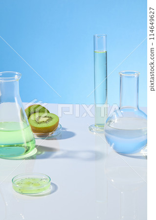 Baby blue background against some laboratory instrument filled by transparent liquid placed on white counter, decorated by green color of kiwi. Photo of front view, vacant space for advertising 114649627
