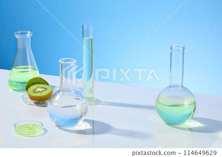 Photograph with high angle shot over light blue background, various types of laboratory glassware and kiwi fruit displayed on white table top. Empty space in center for showing product Photograph with high angle shot over light blue background, various types of laboratory glassware and kiwi fruit displayed on white table top. Empty space in center for showing product 114649629