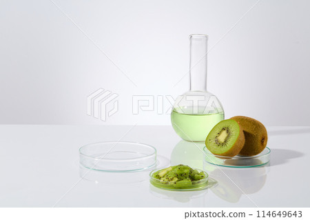 Minimalist style photo on white texture, some dishes containing fresh and chopped kiwi, a boiling flask filled by light green liquid next to an empty petri dish. Displaying space, front view 114649643