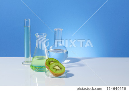 Frontal shot photography over blue background, measuring cylinder, boiling and erlenmeyer flask filled by transparent liquid, kiwi pieces decorated in front. Blank space for designing, copy space Frontal shot photography over blue background, measuring cylinder, boiling and erlenmeyer flask filled by transparent liquid, kiwi pieces decorated in front. Blank space for designing, copy space 114649656