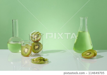 Beakers containing kiwi cut in halves. Few flasks filled with green liquid extracted from kiwi. Empty space in the middle for cosmetic product presentation with laboratory concept Beakers containing kiwi cut in halves. Few flasks filled with green liquid extracted from kiwi. Empty space in the middle for cosmetic product presentation with laboratory concept 114649705