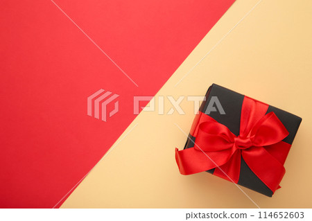 Black gift box with red ribbon and bow on beige-red background. 114652603