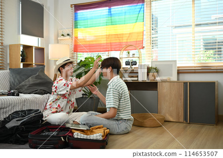 Happy gay couple sitting in living room packing bags, preparing things for vacations trip Happy gay couple sitting in living room packing bags, preparing things for vacations trip 114653507