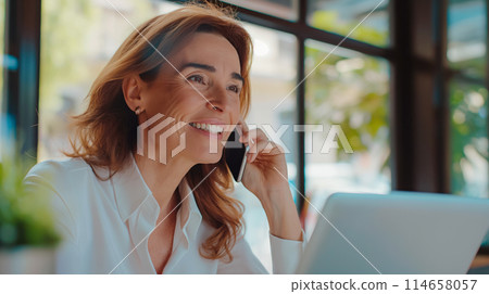 Portrait of a beautiful smiling adult woman talking on the phone, European businesswoman working as a freelancer, laptop standing on the table in front of her, copy space for concept 114658057