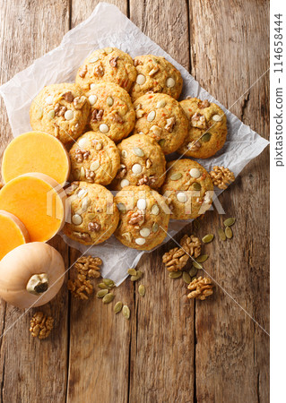 Pumpkin cookies with white chocolate chips, walnut and pumpkin seeds closeup on the parchment. Vertical top view Pumpkin cookies with white chocolate chips, walnut and pumpkin seeds closeup on the parchment. Vertical top view 114658444