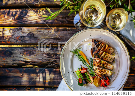 top view of grilled tuna steak with herbs and vegetables, a glass of white wine on a wooden table in a cafe under an olive tree, a trip to the Greek islands and lunch in an old fisherman tavern 114659115