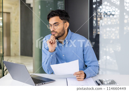 Business professional in blue shirt working at desk with laptop, document, and tablet in modern office environment. 114660449