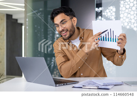 A cheerful man in glasses presenting business charts during a video call from a modern office, explaining growth trends to colleagues. 114660548