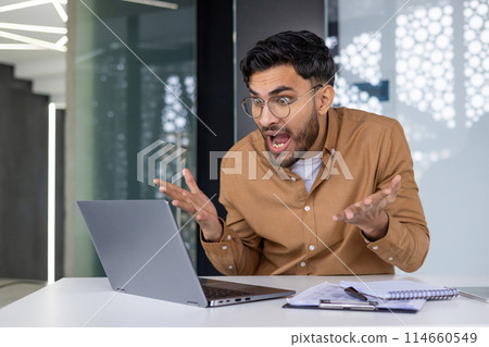 Shocked young man with glasses and beard, gesturing, looking at laptop in modern office. Emotional reaction to unexpected news or work situation. Shocked young man with glasses and beard, gesturing, looking at laptop in modern office. Emotional reaction to unexpected news or work situation. 114660549
