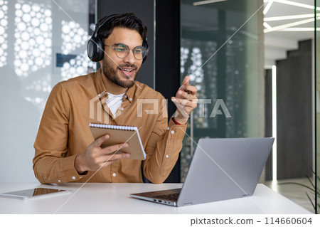 Man wearing headphones attending an online webinar while taking notes. He is sitting at a modern office desk with a laptop and notebook. Man wearing headphones attending an online webinar while taking notes. He is sitting at a modern office desk with a laptop and notebook. 114660604