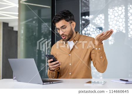 Office worker looking frustrated as he reads bad news on his phone while working on his laptop. 114660645