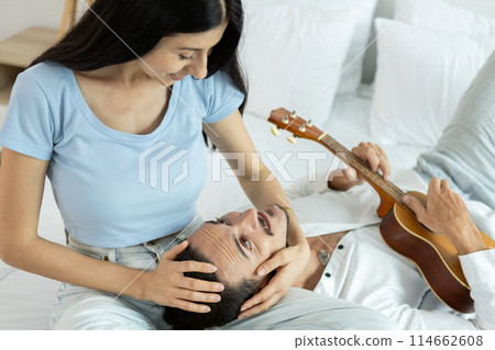 happy couple playing ukulele on bed in bedroom. man and woman enjoying playing music creates happy 114662608