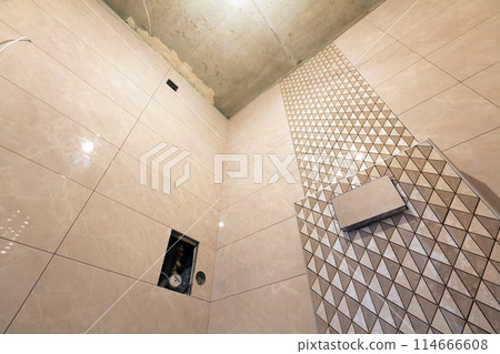 Unfinished reconstruction of bathroom or toilet with light beige geometric mosaic ceramic tiles installed on walls, place for lavatory and bath. Low angle, bottom view. Unfinished reconstruction of bathroom or toilet with light beige geometric mosaic ceramic tiles installed on walls, place for lavatory and bath. Low angle, bottom view. 114666608