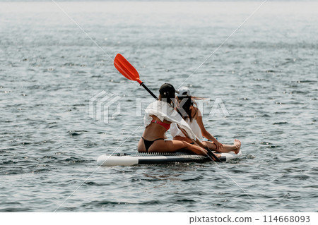 Two women floating on a surfboard in the ocean. One of them is wearing a white shirt and the other is wearing a black bikini. Two women floating on a surfboard in the ocean. One of them is wearing a white shirt and the other is wearing a black bikini. 114668093