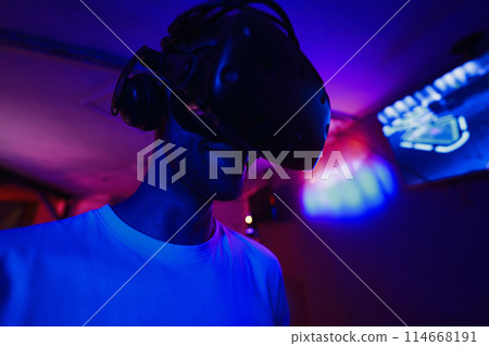 A player plays a game using a virtual reality helmet in a neon room. Close-up photo. A player plays a game using a virtual reality helmet in a neon room. Close-up photo. 114668191