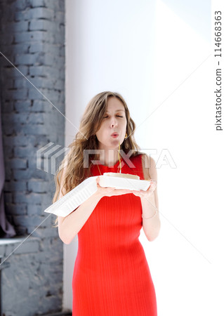 Beautiful and young girl with long hair in a red dress blows out a candle on a bento cake at a party Beautiful and young girl with long hair in a red dress blows out a candle on a bento cake at a party 114668363