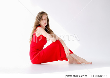 Beautiful and young girl with long hair in a red dress sits on the floor of a cyclorama and poses 114668365