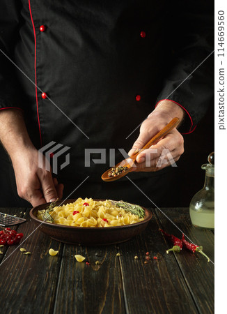Adding dry pepper with a spoon in the cook hand to a plate of pasta. Chef at the kitchen table in a public house preparing a delicious lunch Adding dry pepper with a spoon in the cook hand to a plate of pasta. Chef at the kitchen table in a public house preparing a delicious lunch 114669560