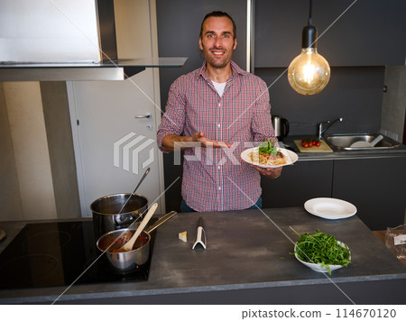 Attractive smiling young man 40s, showing at camera a plate with freshly cooked Italian pasta, standing at kitchen table with fresh ingredients and kitchen utensils. People. Italian food and culture 114670120
