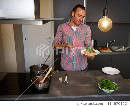 Handsome young Italian chef, man showing a white plate with Italian spaghetti capellini, standing in the home kitchen. Chef showing a served dish with Italian pasta. Italy. Food. Culture. Traditions Handsome young Italian chef, man showing a white plate with Italian spaghetti capellini, standing in the home kitchen. Chef showing a served dish with Italian pasta. Italy. Food. Culture. Traditions 114670122