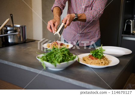 Close-up man cooking dinner in stylish modern minimalist home kitchen interior, grating parmesan cheese over a plate with freshly boiled pasta with tomato sauce. Ingredients on the kitchen countertop 114670127