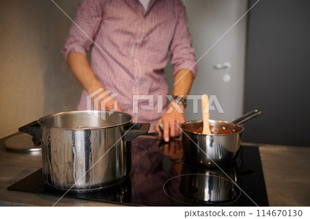 Details on steel saucepan on the induction oven, electric stove against blurred male chef switching on panel for boiling water. Cropped view of blurred man switching on, turning on the electric stove 114670130