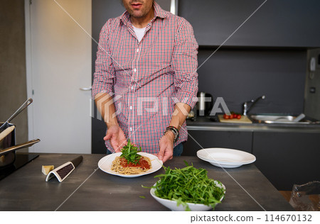 Young man cooking Italian pasta spaghetti for dinner in the home kitchen. Focus on served plate with Italian spaghetti with tomato sauce and greens arugula herbs. People, food and drink consumerism Young man cooking Italian pasta spaghetti for dinner in the home kitchen. Focus on served plate with Italian spaghetti with tomato sauce and greens arugula herbs. People, food and drink consumerism 114670132