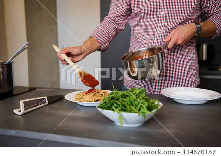 Close-up chef pouring tomato sauce on Italian pasta, plating up the dish before serving spaghetti. A bunch of organic cherry tomato on foreground. Man cooking dinner at home. Cuisine. Culinary. Food 114670137