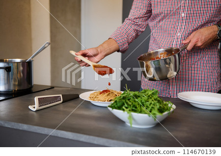 Handsome young man pouring tomato sauce over freshly boiled pasta, plating up the dish, standing at kitchen counter with fresh organic ingredients on the kitchen countertop 114670139