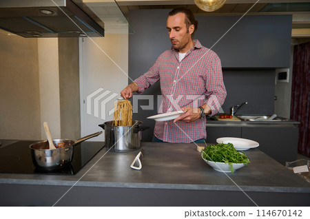 Handsome young male chef pouring freshly cooked Italian past into a plate, ready to serve it to the customers. Food and drink consumerism. Culinary. Epicure. Man cooking Italian spaghetti for dinner 114670142