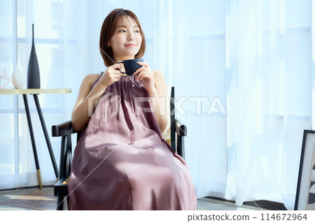 A woman sitting on a chair in a room and drinking coffee 114672964
