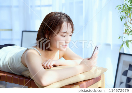 一位女士躺在房間的椅子上，看著她的智慧型手機 114672979